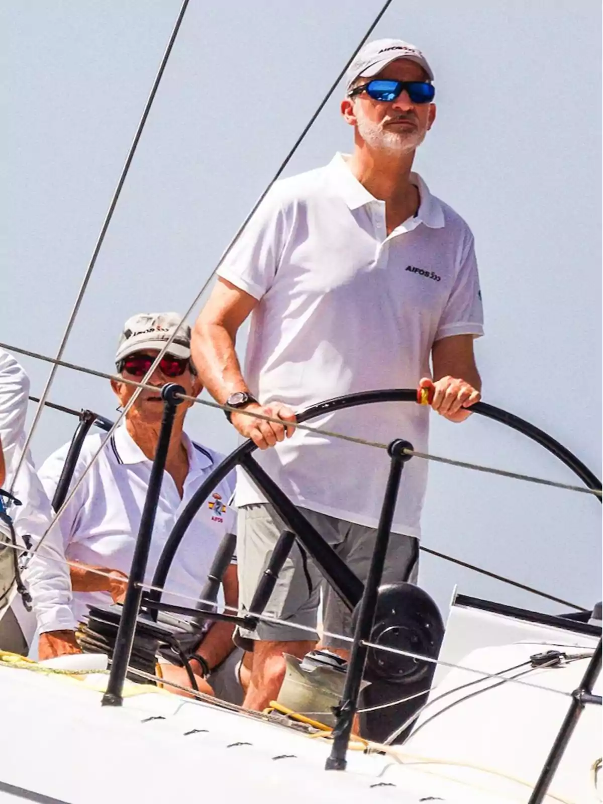 Personas navegando en un velero, una de ellas está al timón y lleva gafas de sol y gorra.