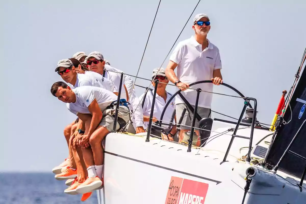 Un grupo de personas con ropa deportiva blanca y gorras grises navegando en un velero.