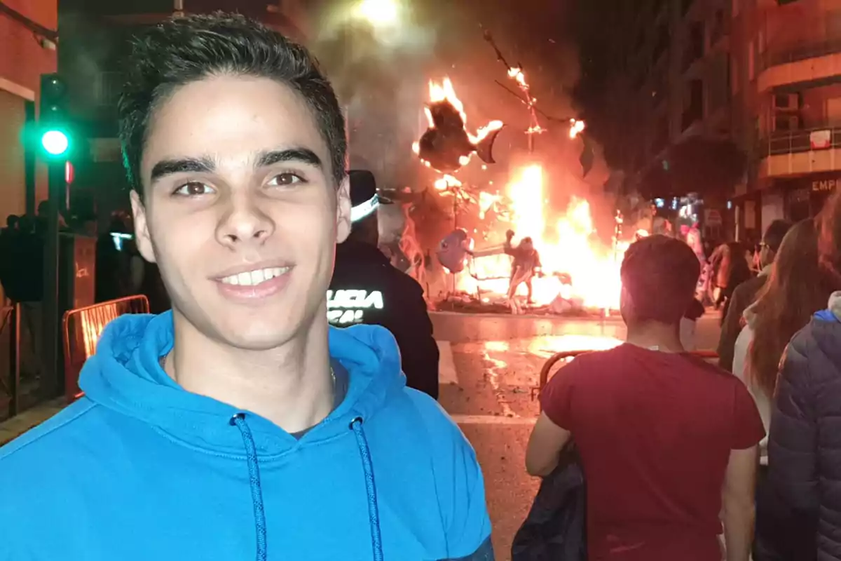 Un joven con sudadera azul sonríe frente a una hoguera en una calle concurrida durante la noche.