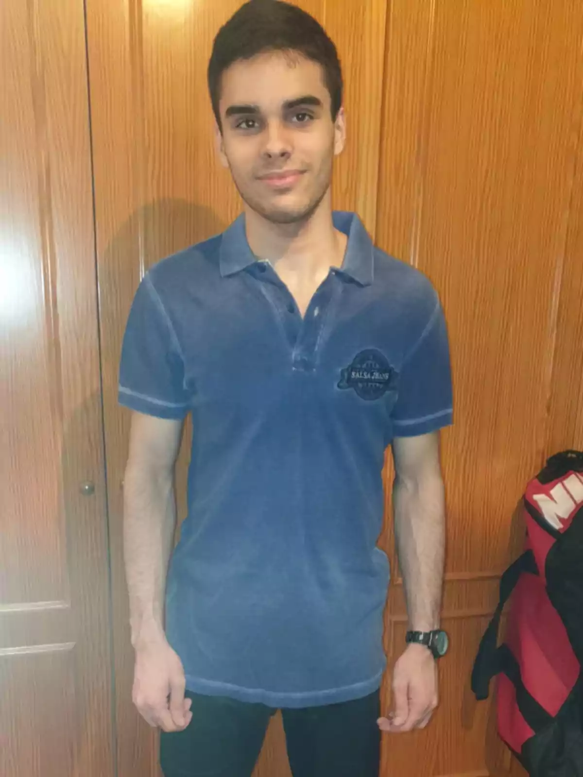 Un joven con una camiseta azul de pie frente a un armario de madera.