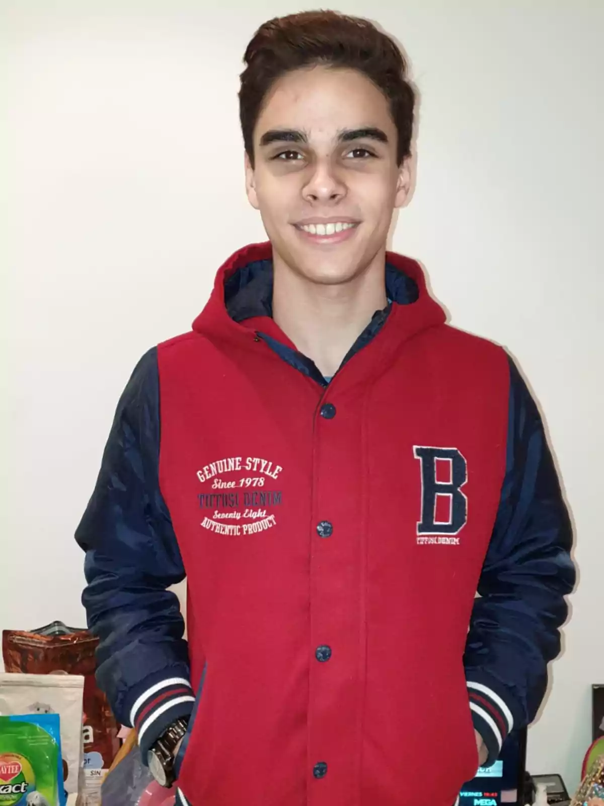 Un joven sonriente con una chaqueta roja y azul con la letra 