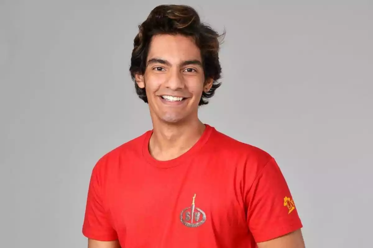 Un hombre joven con cabello oscuro y ondulado, sonriendo y vistiendo una camiseta roja con un logotipo en el pecho y una figura en la manga.