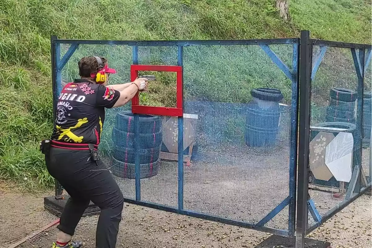 Persona participando en una competencia de tiro al blanco, usando protección auditiva y visual, apuntando con una pistola a través de una ventana en una estructura de malla azul con un marco rojo.