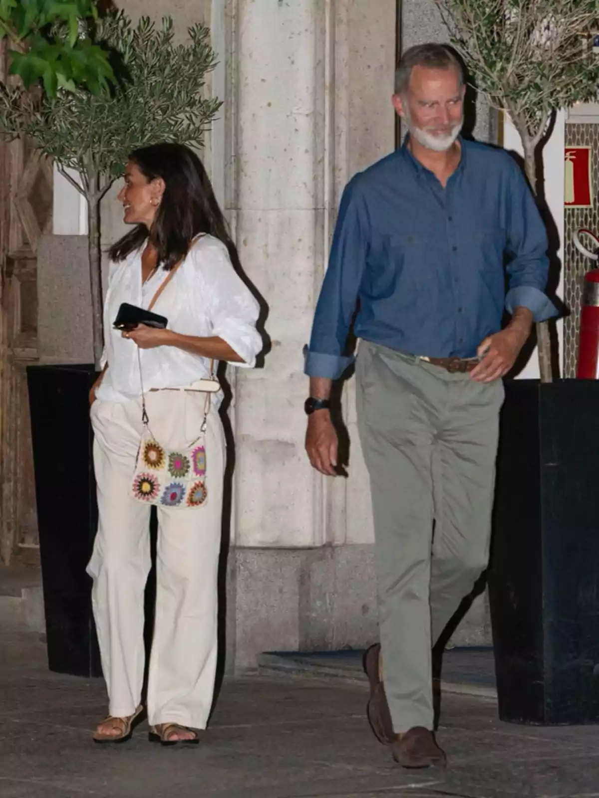 Una mujer y un hombre caminando juntos en una calle, ella lleva una blusa blanca y pantalones claros, mientras que él viste una camisa azul y pantalones verdes.