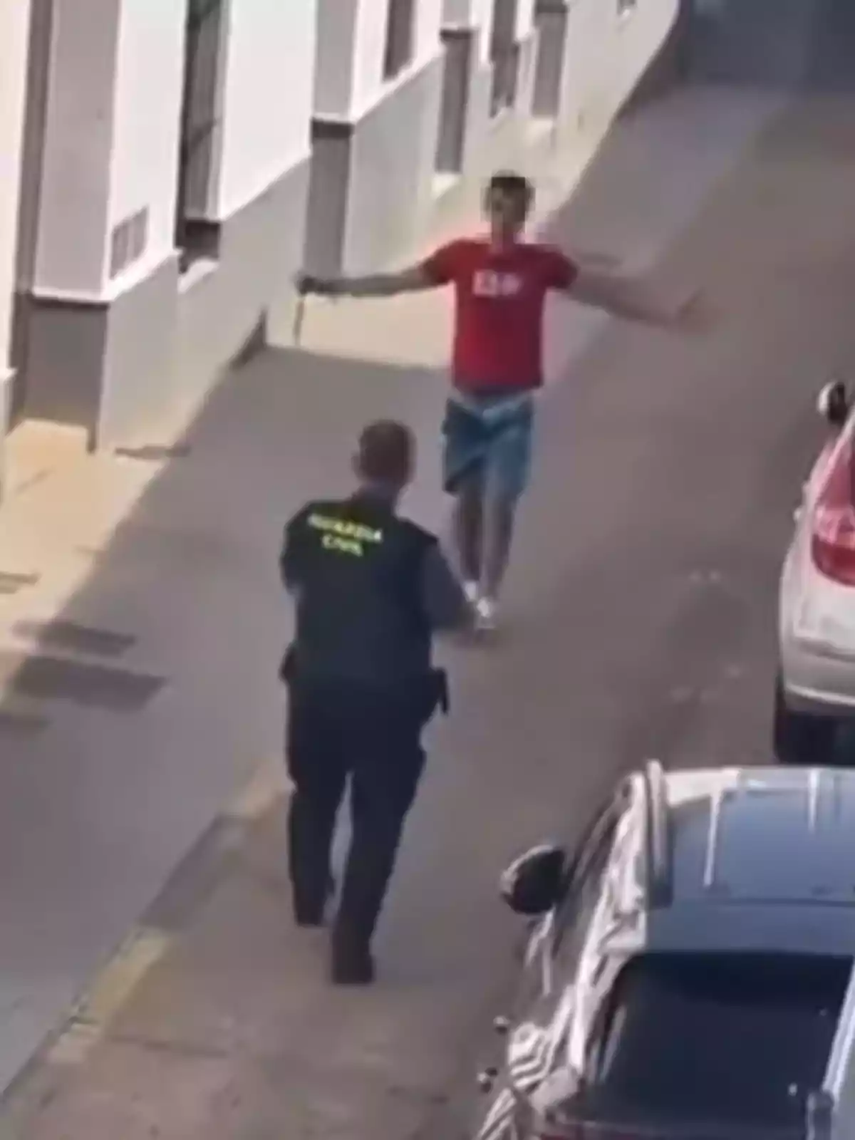 Un agente de la Guardia Civil se aproxima a un hombre que lleva una camiseta roja y pantalones cortos de mezclilla en una calle estrecha.