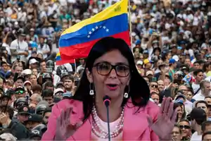 Una mujer con gafas y chaqueta rosa habla frente a un micrófono con una multitud y una bandera de Venezuela de fondo.