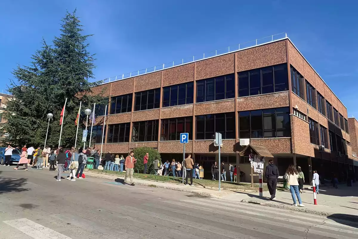 Personas reunidas frente a un edificio de ladrillo con ventanas grandes y varias banderas ondeando, en un día soleado.