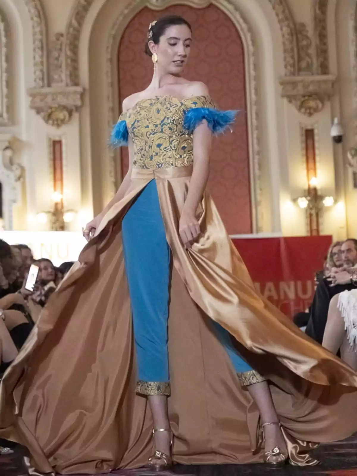 Una modelo vestida por Manu Sánchez camina por la pasarela luciendo un vestido elegante con detalles dorados y azules, en un entorno de arquitectura clásica y público observando.