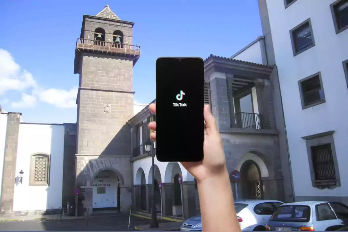 Una mano sostiene un teléfono con la aplicación TikTok abierta frente a un edificio histórico con una torre y un cielo azul.