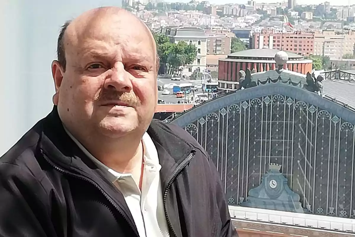 Un hombre con bigote y chaqueta oscura está frente a un edificio con una estructura de vidrio y metal en una ciudad.