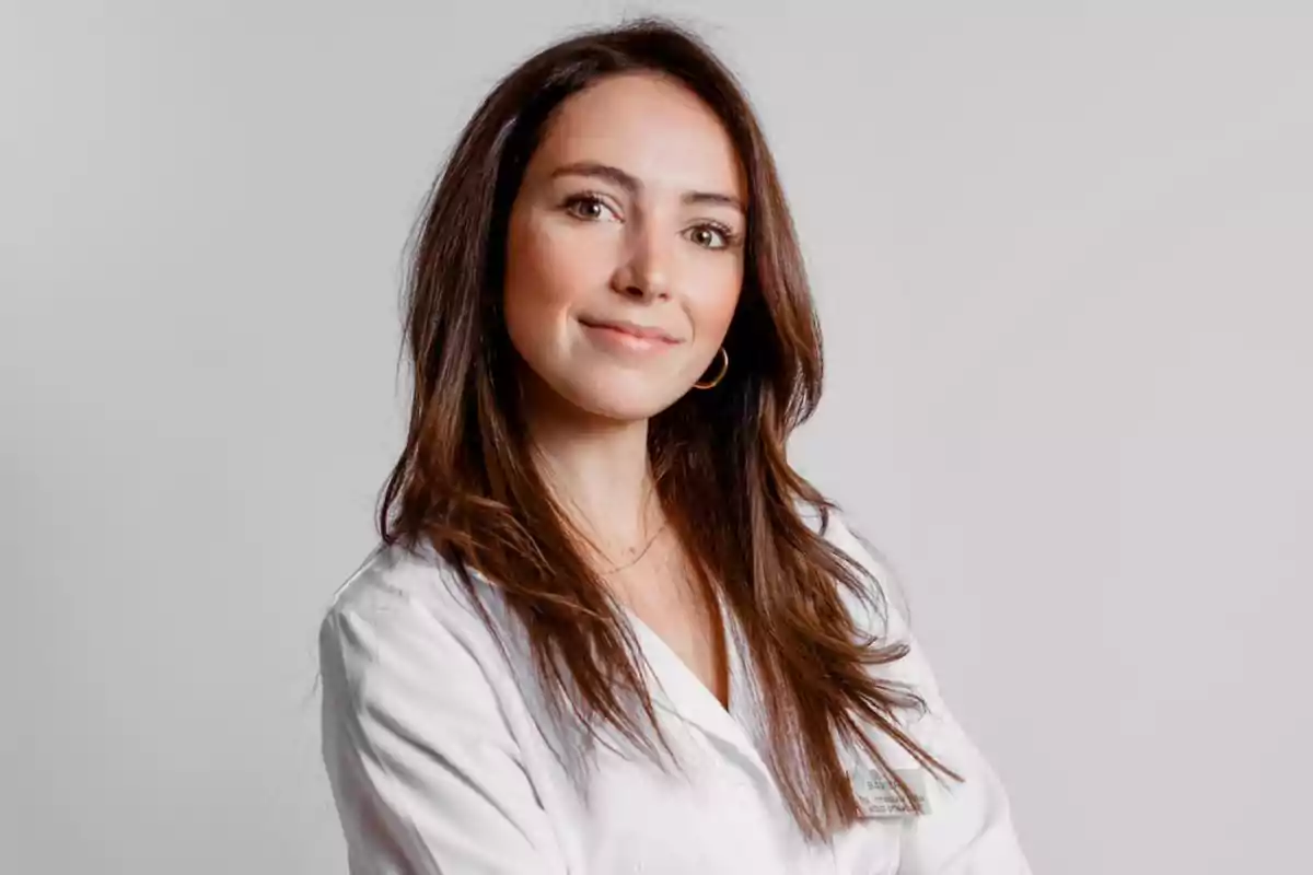 Mujer con bata blanca sonriendo frente a un fondo gris.