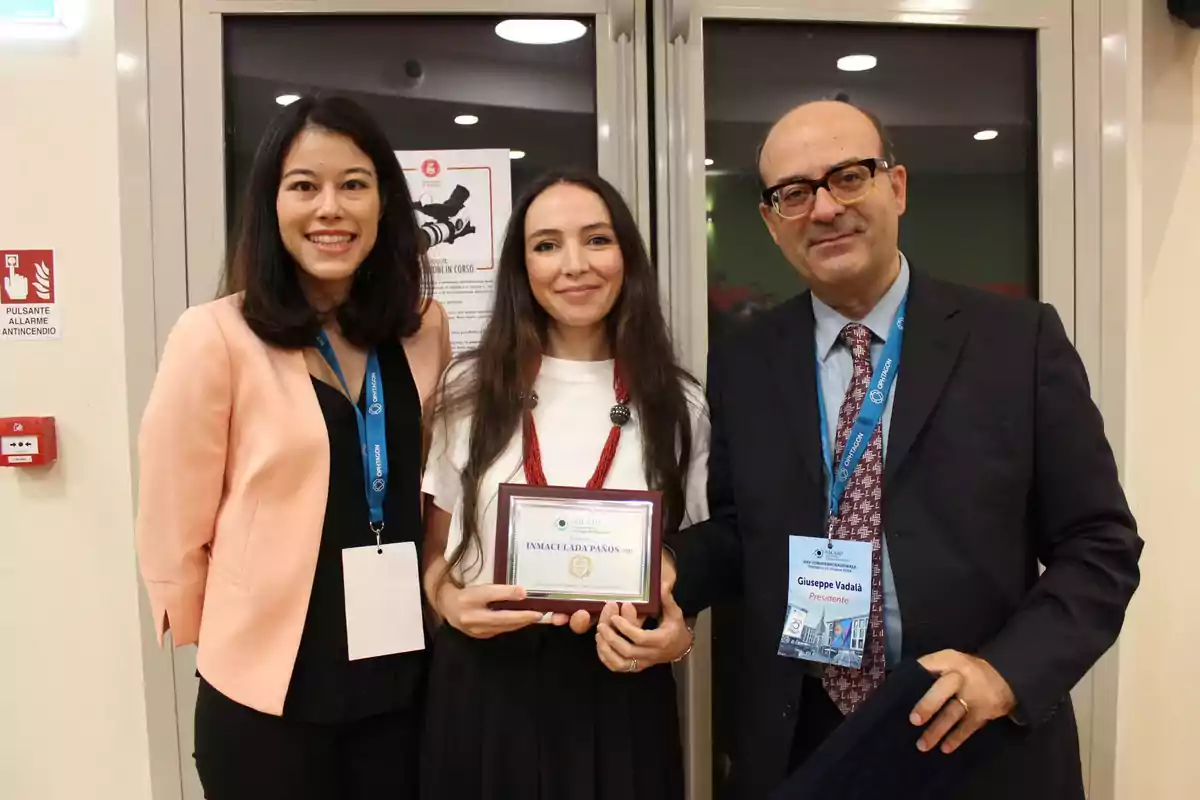 Tres personas posan para una foto en un evento, una de ellas sostiene un reconocimiento enmarcado.