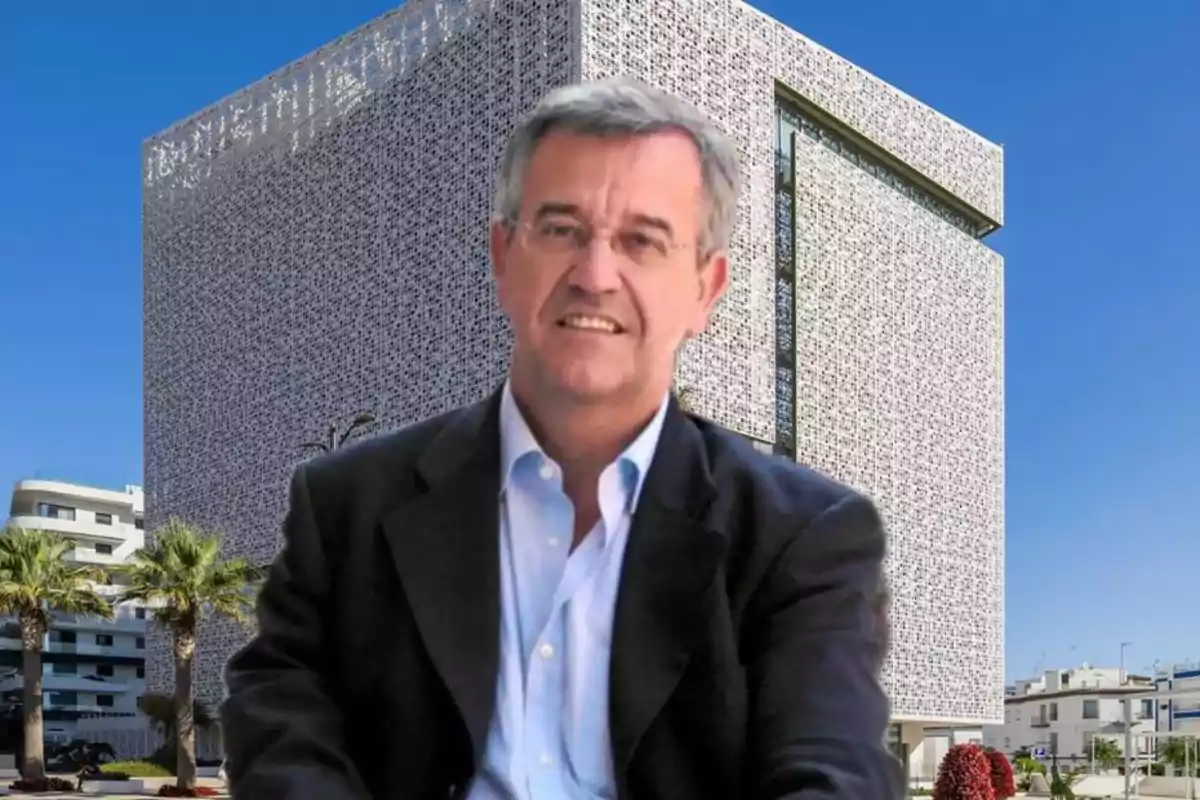 Un hombre con gafas y traje oscuro está frente a un edificio moderno con una fachada decorativa y cielo azul.