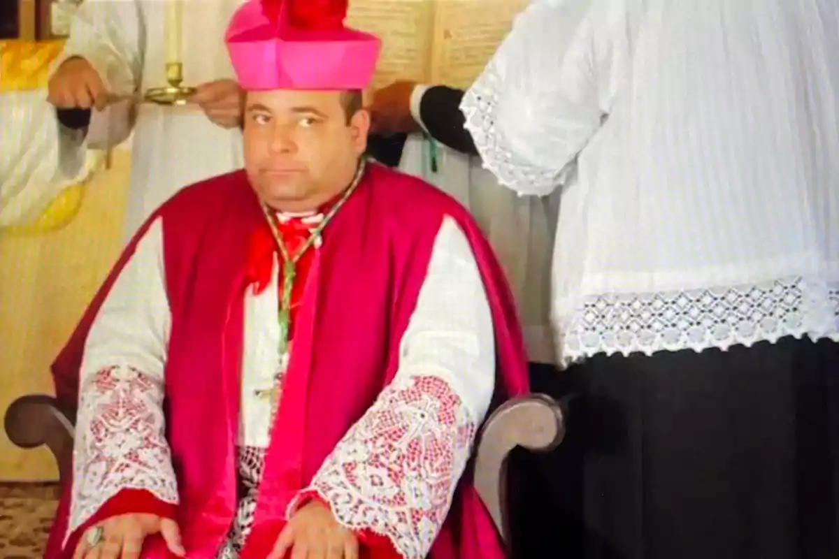 Un hombre vestido con vestimenta eclesiástica de color rojo y rosa, sentado en una silla, con una expresión seria, mientras otra persona de pie a su lado sostiene un libro abierto.