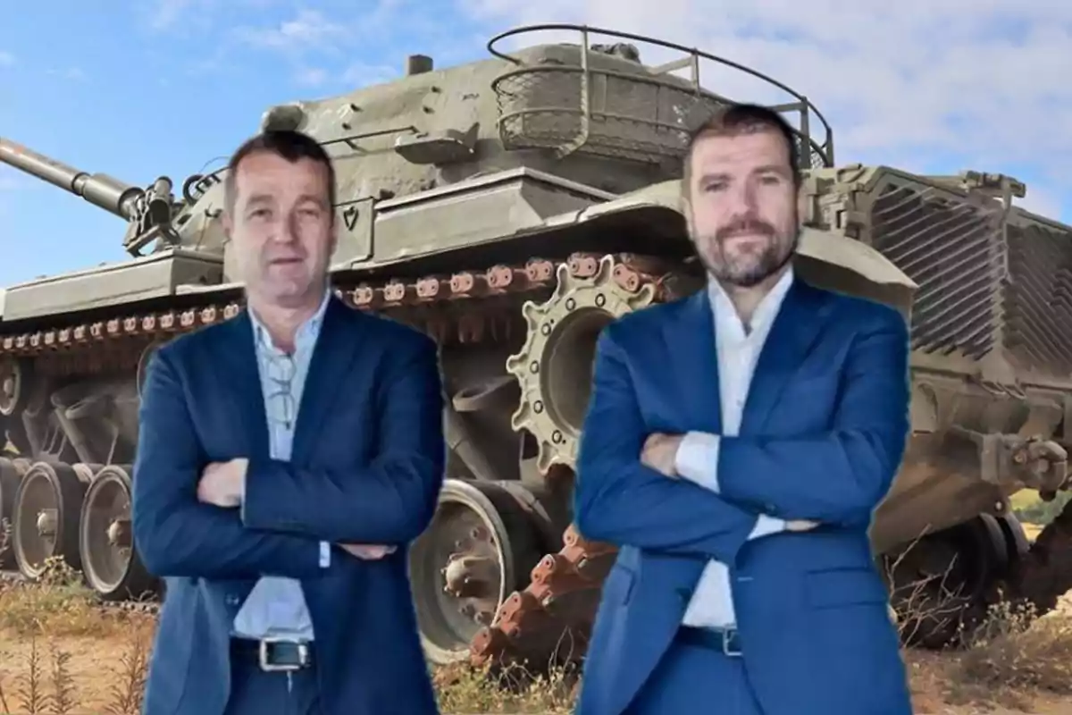 Dos hombres de traje posan con los brazos cruzados frente a un tanque militar en un entorno al aire libre.