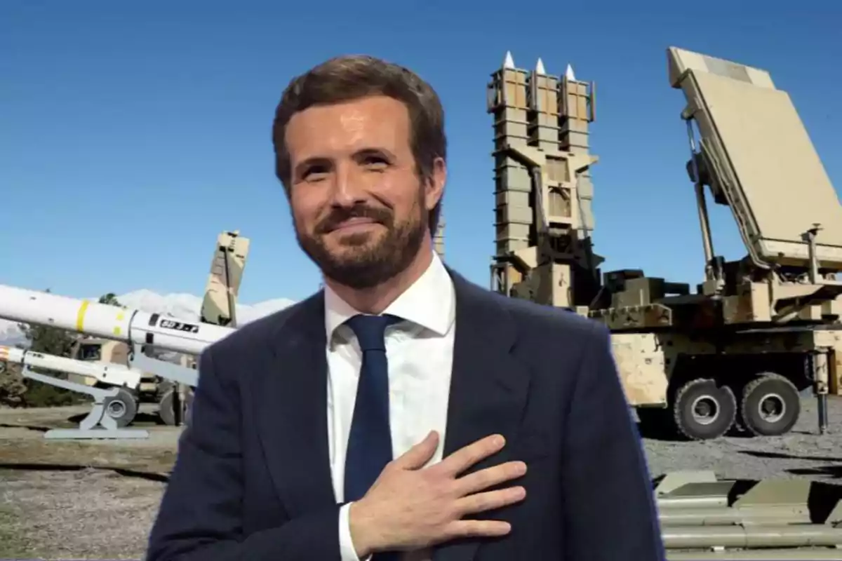 Un hombre con traje y corbata sonríe frente a un fondo de equipo militar y cielo despejado.