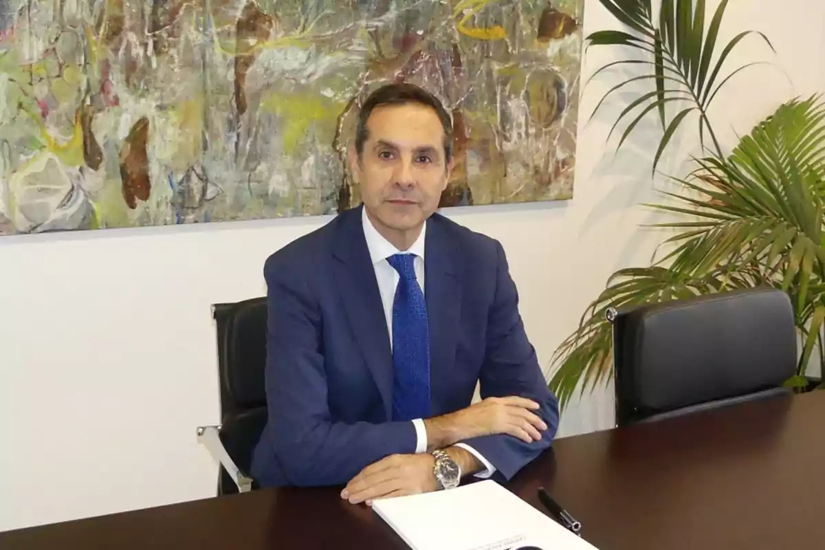 Un hombre con traje azul está sentado en una mesa con un cuaderno y un bolígrafo, detrás hay una pintura abstracta y una planta.