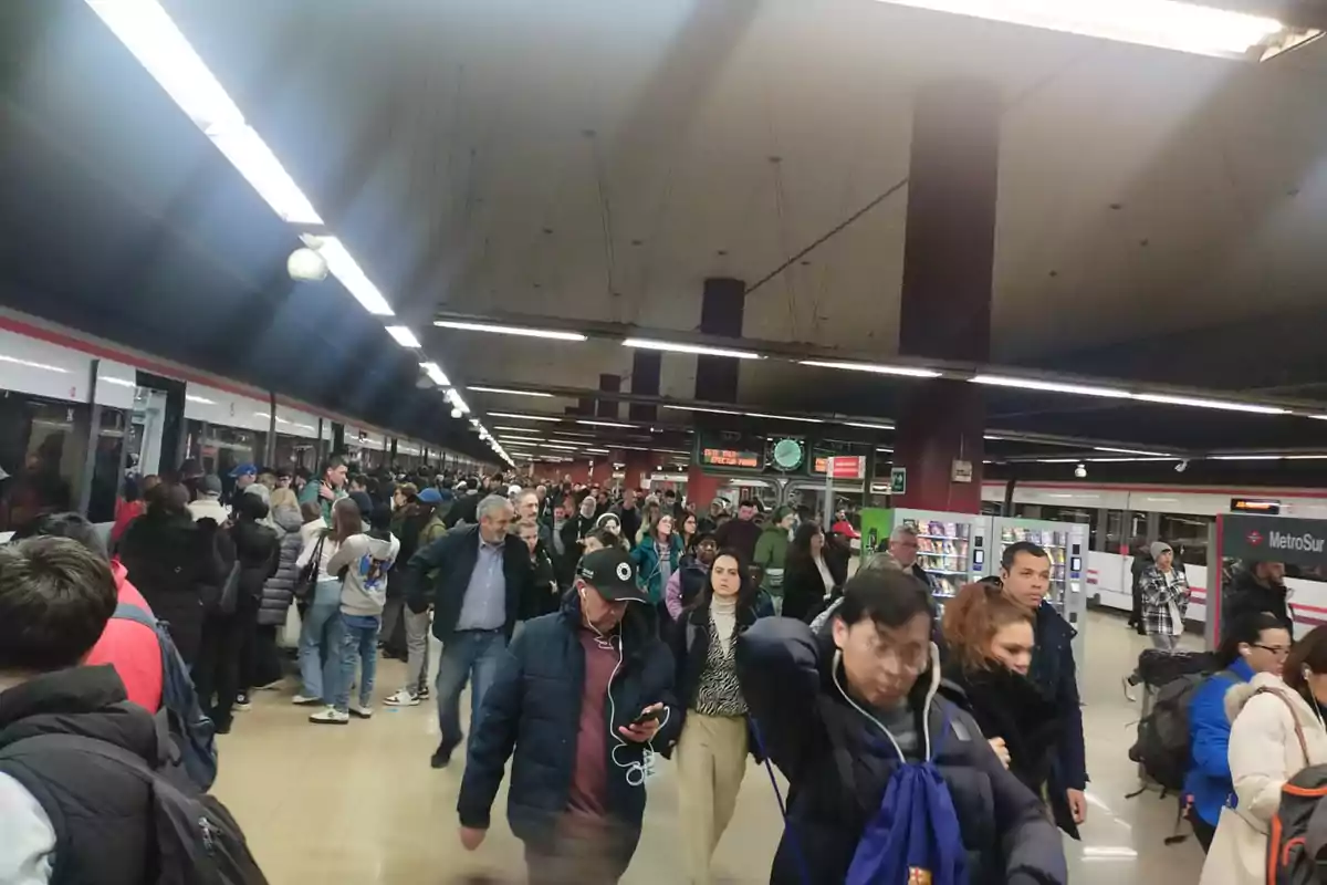 Una multitud de personas camina por un andén de metro concurrido, con un tren detenido a la izquierda y varias tiendas al fondo.