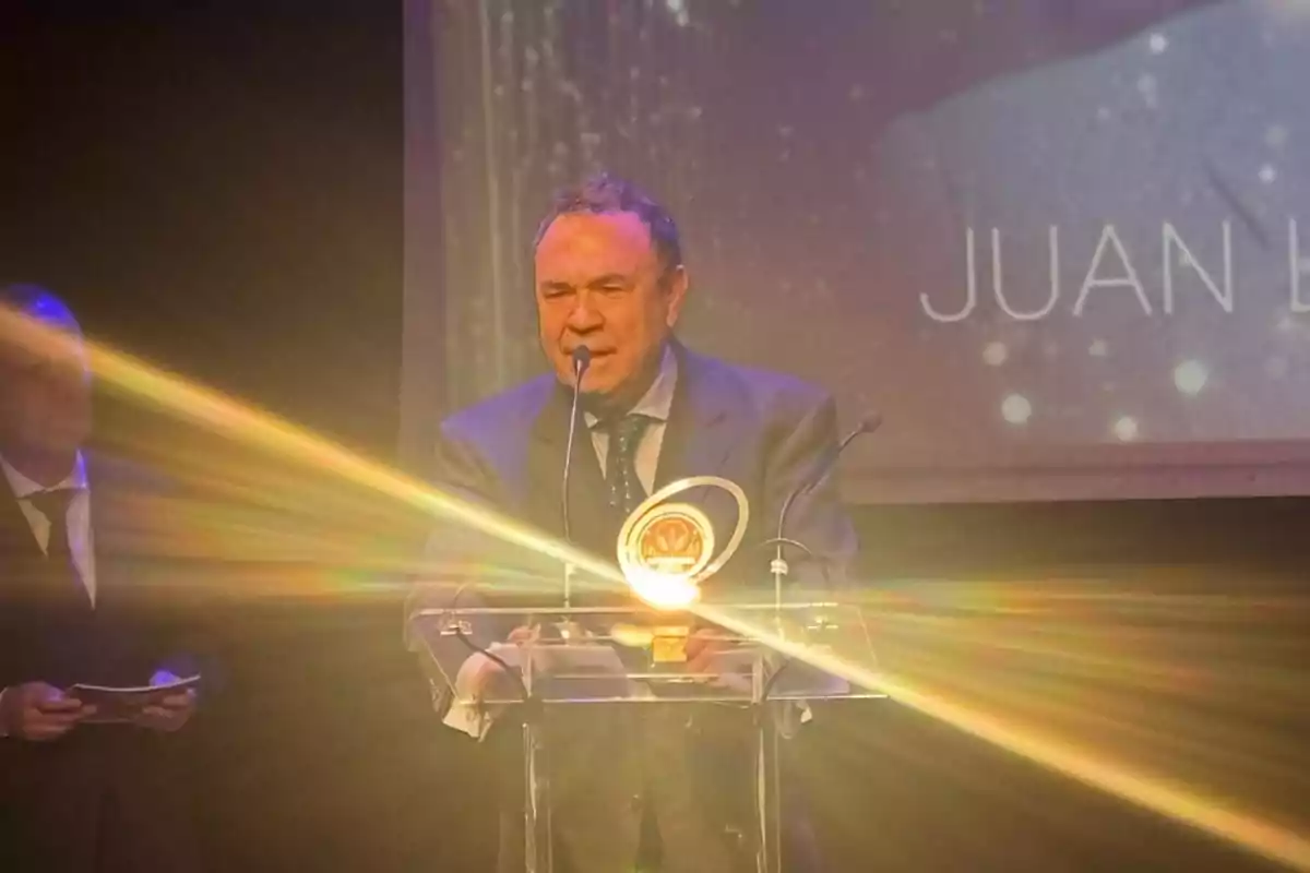 Un hombre en un escenario hablando al micrófono con un trofeo iluminado frente a él.