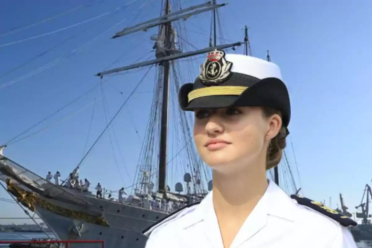 Una mujer con uniforme naval y sombrero de oficial está de pie frente a un gran velero con cielo despejado de fondo.