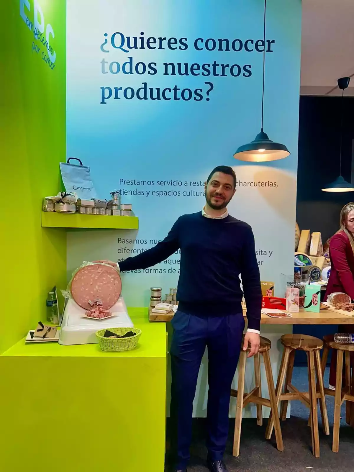Un hombre de pie junto a un mostrador con productos alimenticios y un cartel que invita a conocer más sobre ellos. Un hombre de pie junto a un mostrador con productos alimenticios y un cartel que invita a conocer más sobre ellos.