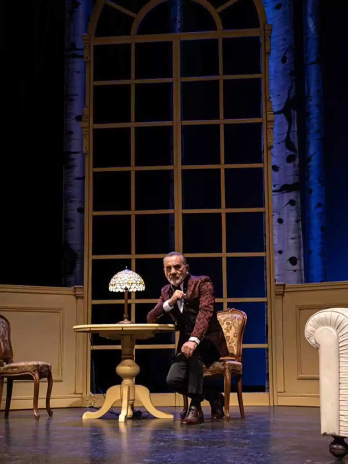 Un hombre posa junto a una mesa con una lámpara en un escenario teatral decorado con un gran ventanal y muebles clásicos. Un hombre posa junto a una mesa con una lámpara en un escenario teatral decorado con un gran ventanal y muebles clásicos.