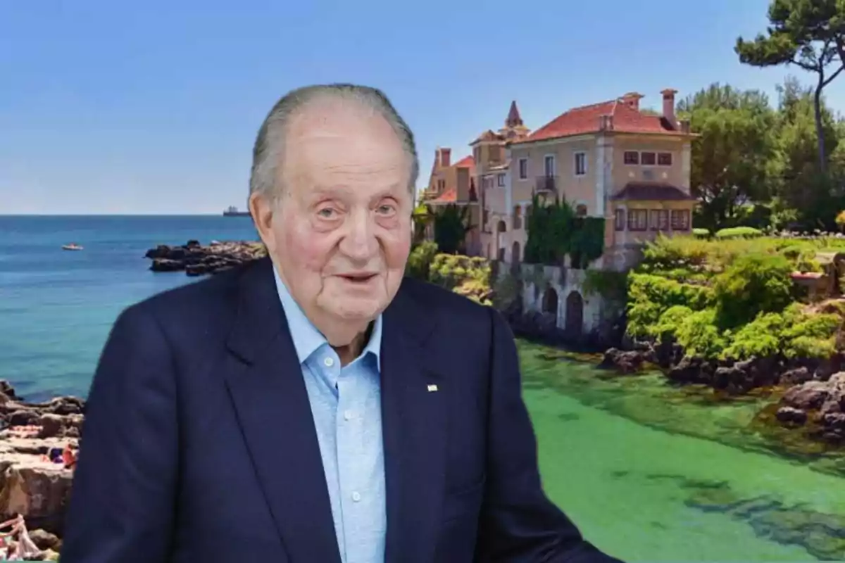 An older man in a dark suit and light blue jersey is in front of a coastal landscape with a large house and trees in the background.