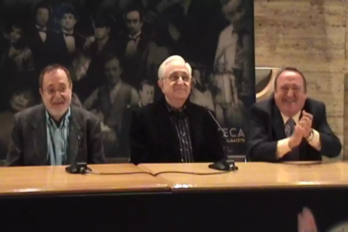Tres hombres sentados detrás de una mesa en una conferencia de prensa, con un fondo de imágenes en blanco y negro de figuras históricas. Tres hombres sentados detrás de una mesa en una conferencia de prensa, con un fondo de imágenes en blanco y negro de figuras históricas.