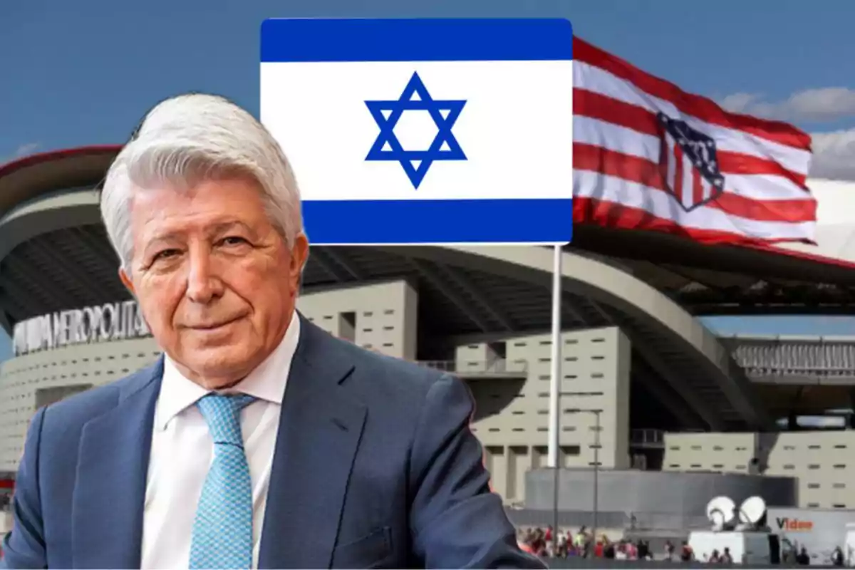 Un hombre de cabello canoso y traje azul está frente a un estadio con una bandera de Israel y otra del Atlético de Madrid ondeando en el fondo.