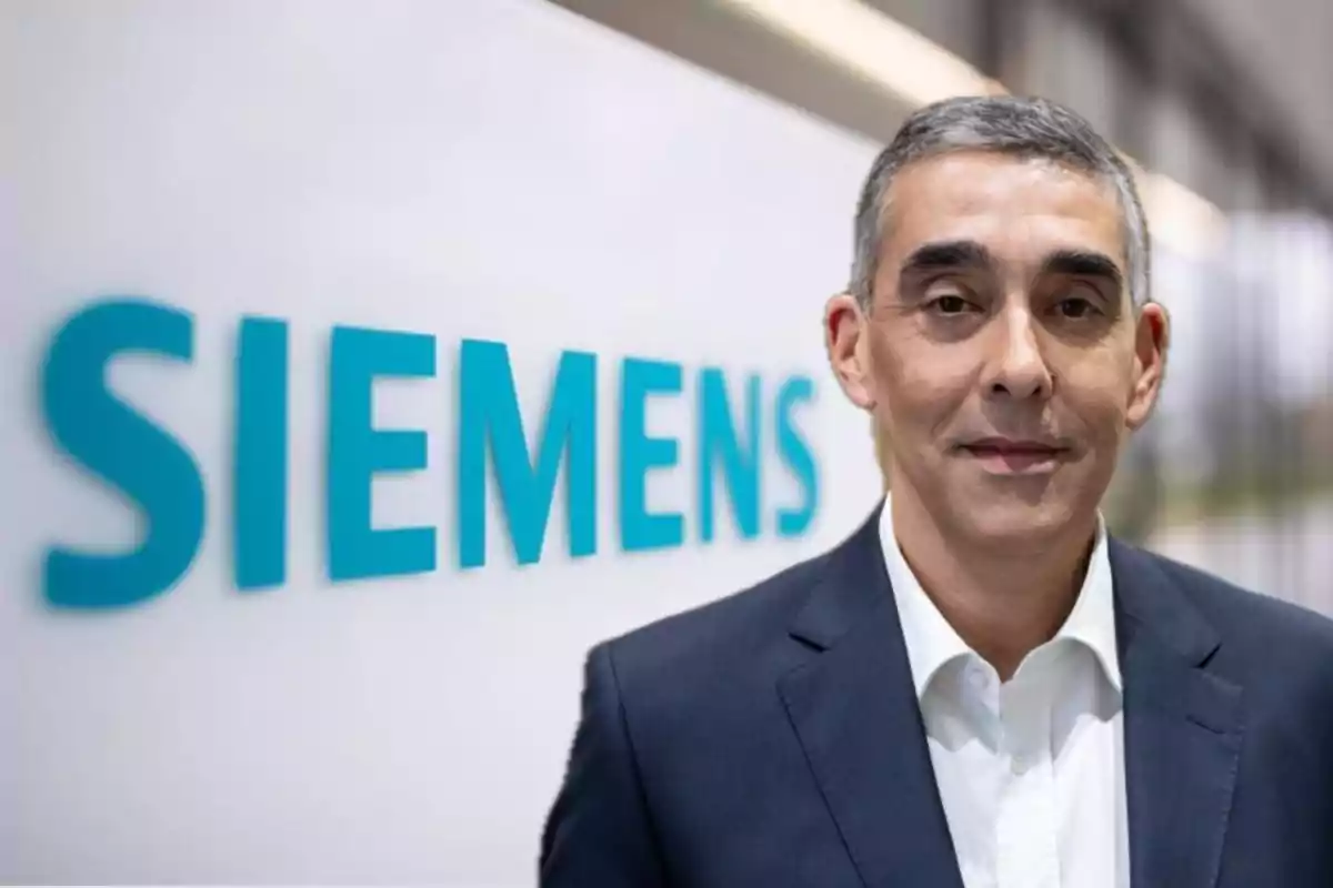 Hombre de cabello canoso con traje oscuro y camisa blanca posando frente a un letrero de Siemens en una oficina moderna