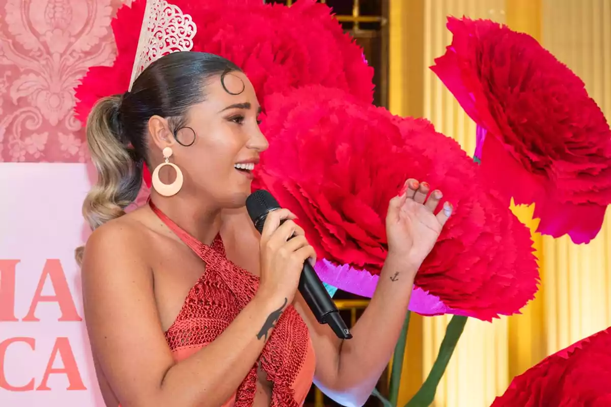 Mujer con vestido rojo y corona blanca hablando por micrófono frente a flores gigantes de papel color fucsia Mujer con vestido rojo y corona blanca hablando por micrófono frente a flores gigantes de papel color fucsia