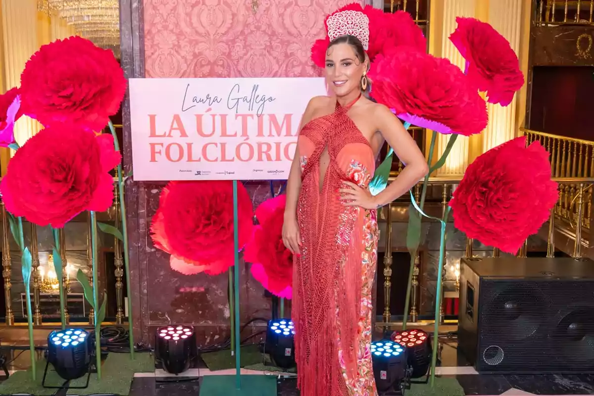 Mujer vestida con un traje rojo y tocado a juego posando sonriente frente a un cartel que dice Laura Gallego La Última Folclórica rodeada de grandes flores rojas decorativas en un ambiente elegante Mujer vestida con un traje rojo y tocado a juego posando sonriente frente a un cartel que dice Laura Gallego La Última Folclórica rodeada de grandes flores rojas decorativas en un ambiente elegante