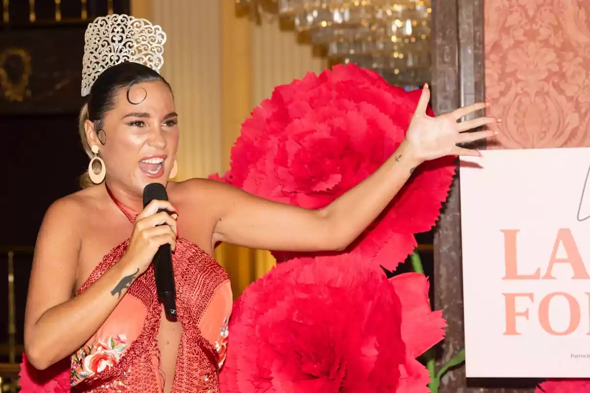 Mujer vestida con traje rojo brillante y tocado blanco cantando con micrófono en mano frente a grandes flores rojas decorativas