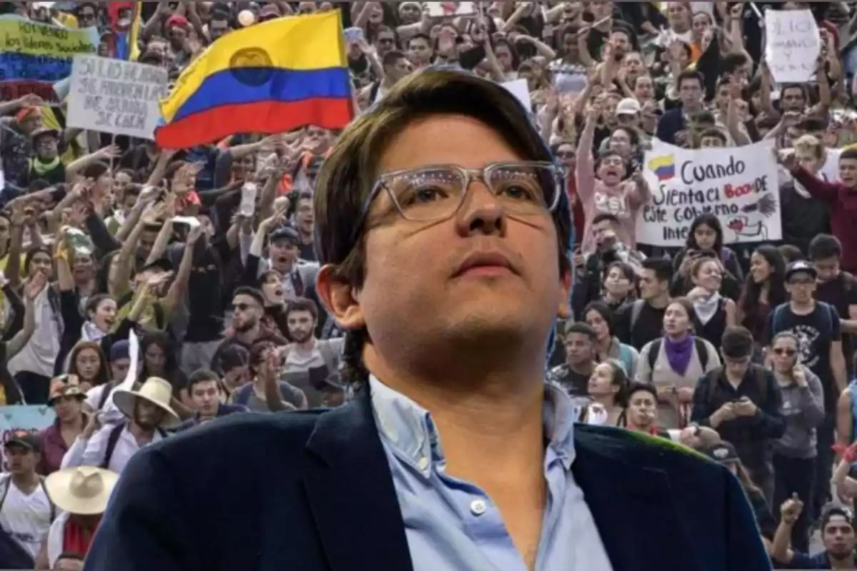 Un hombre con gafas y saco azul aparece en primer plano mientras al fondo se observa una multitud de personas manifestándose con pancartas y una bandera de Colombia.