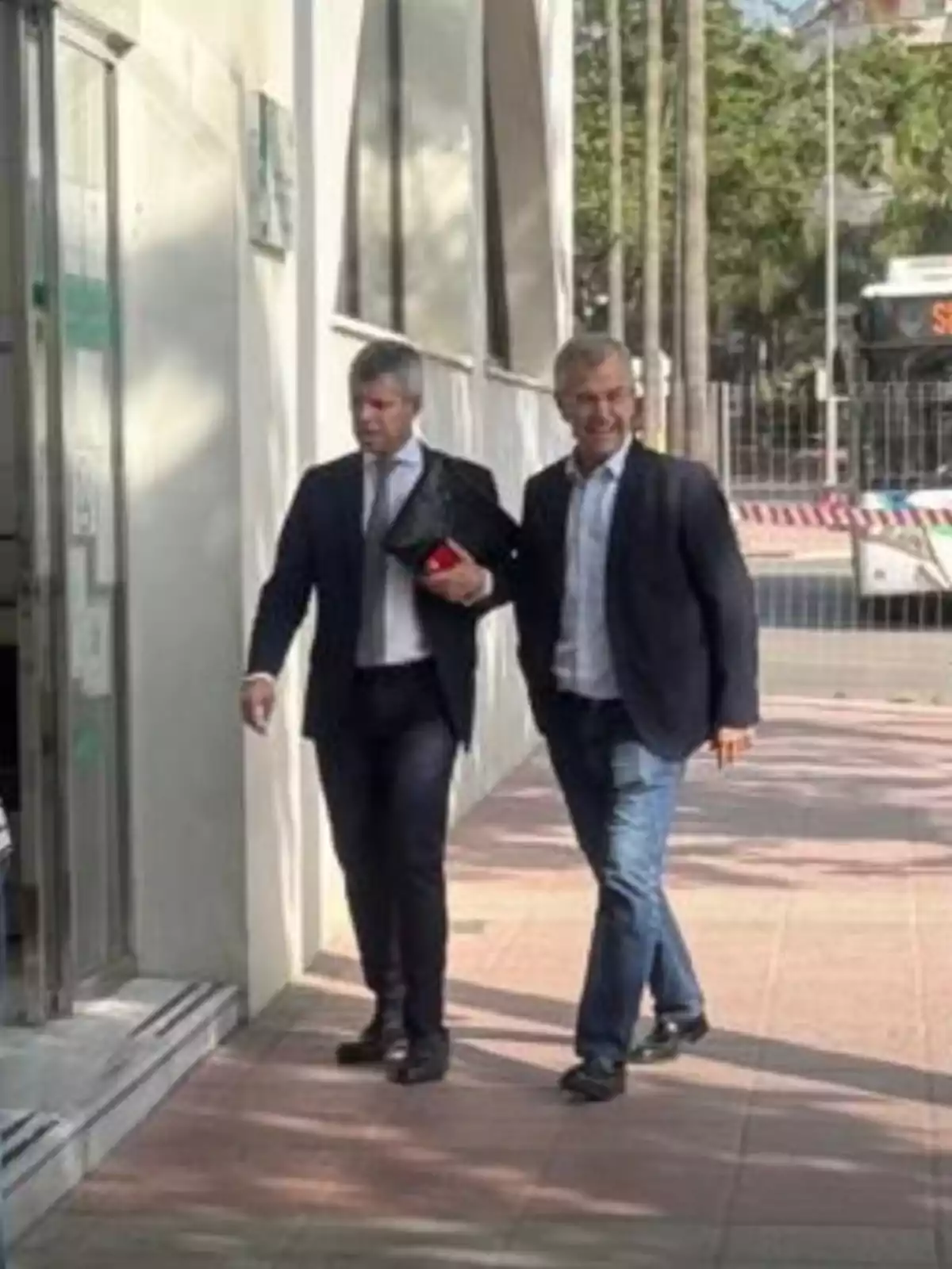 Dos hombres vestidos de traje caminan por una acera junto a un edificio en un entorno urbano y soleado Dos hombres vestidos de traje caminan por una acera junto a un edificio en un entorno urbano y soleado