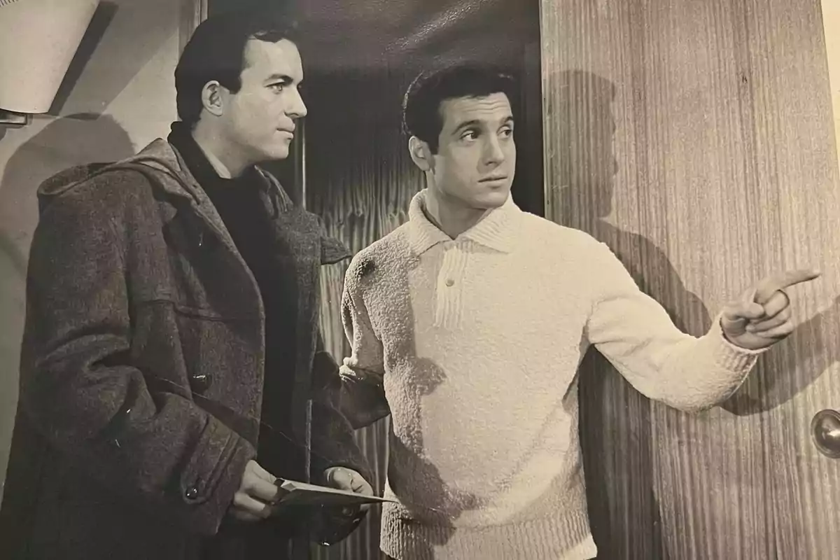 Dos hombres en blanco y negro están de pie junto a una puerta, uno de ellos señala hacia la derecha mientras el otro sostiene unos papeles. Dos hombres en blanco y negro están de pie junto a una puerta, uno de ellos señala hacia la derecha mientras el otro sostiene unos papeles.