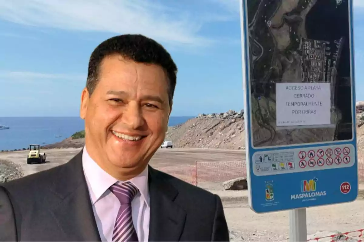 Hombre sonriente con traje y corbata frente a una playa en obras con un cartel que indica el cierre temporal del acceso en Maspalomas