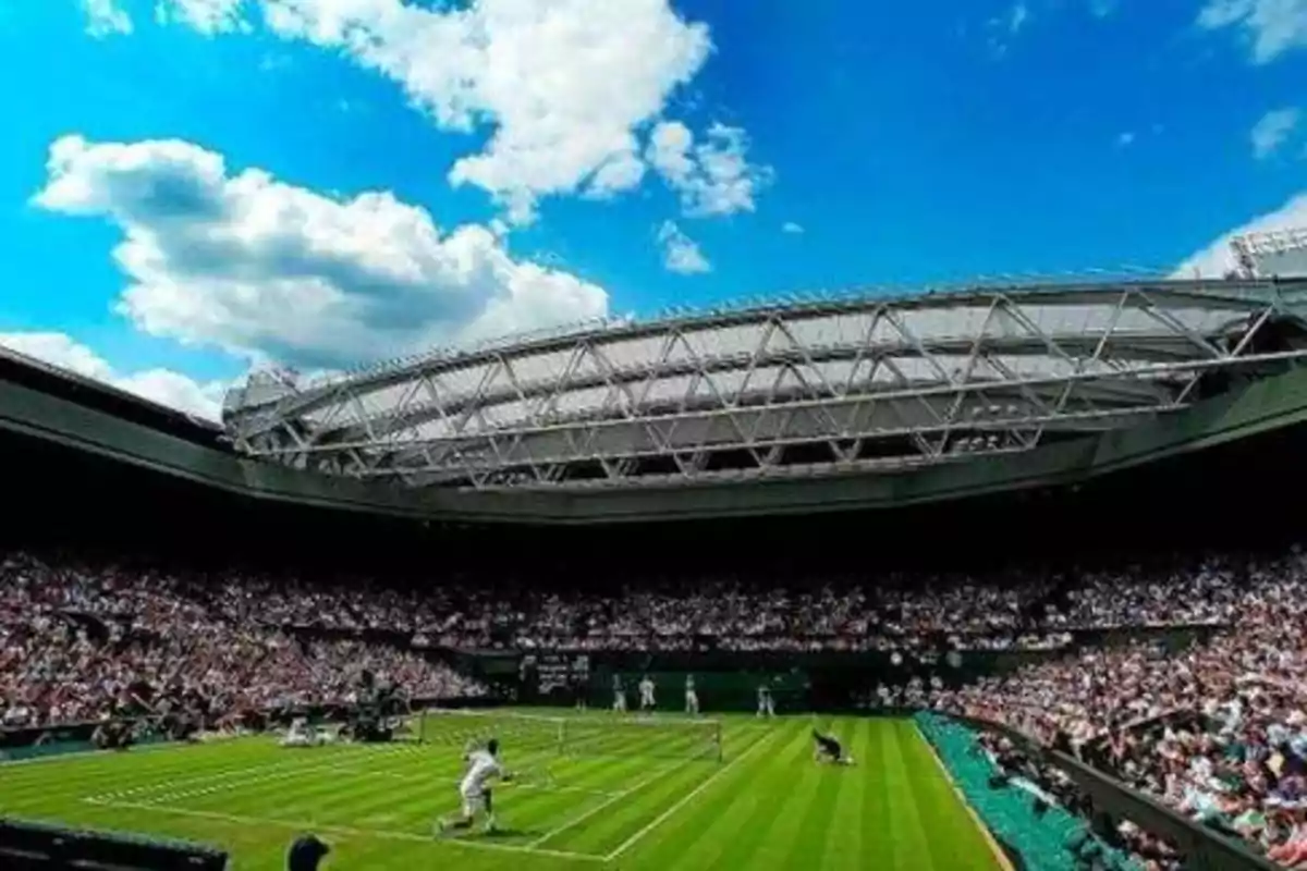 Un partido de tenis en un estadio con techo retráctil y una multitud de espectadores bajo un cielo azul con nubes.