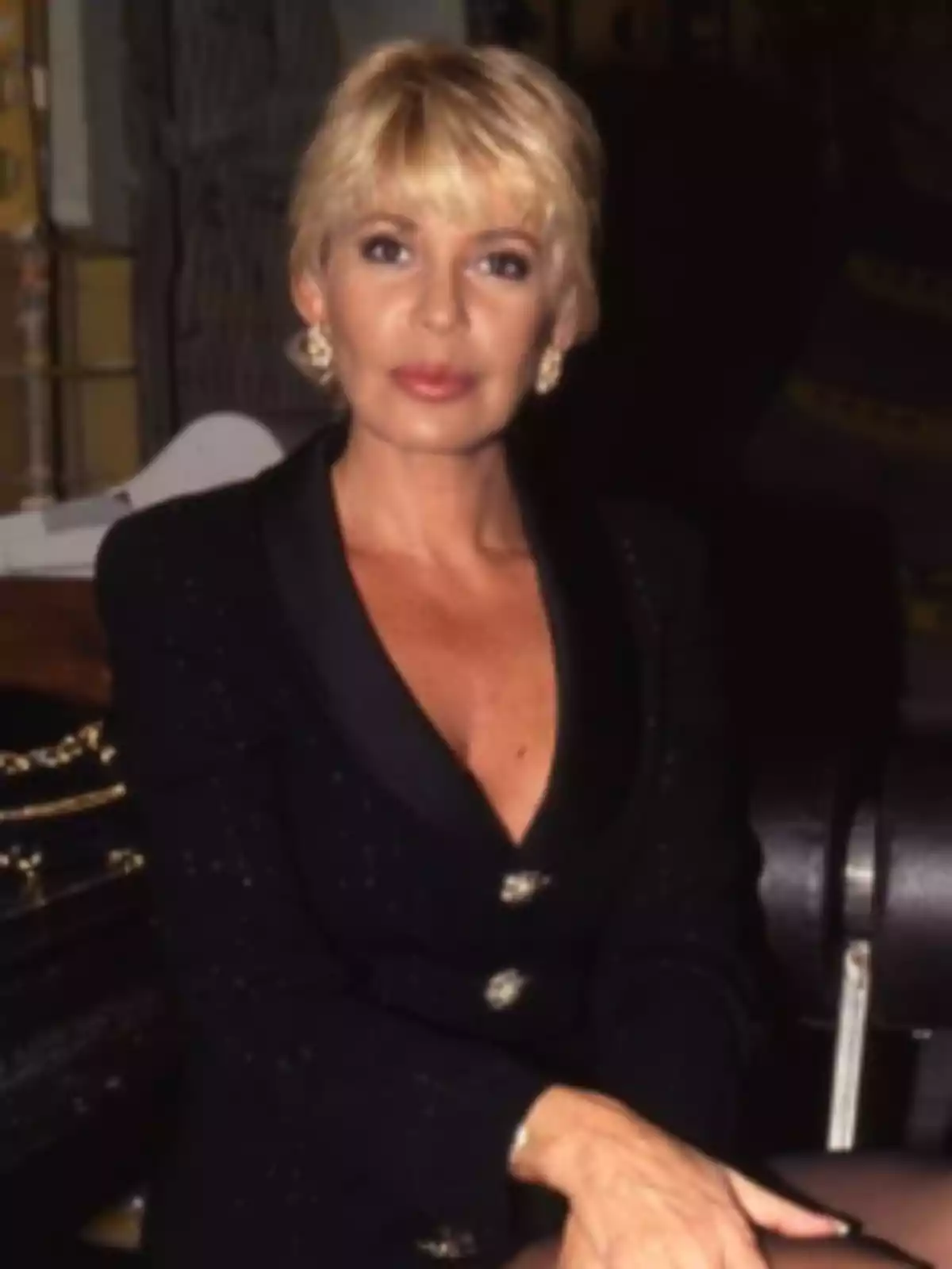 Mujer rubia con cabello corto y vestimenta elegante de color oscuro sentada en un ambiente interior Mujer rubia con cabello corto y vestimenta elegante de color oscuro sentada en un ambiente interior