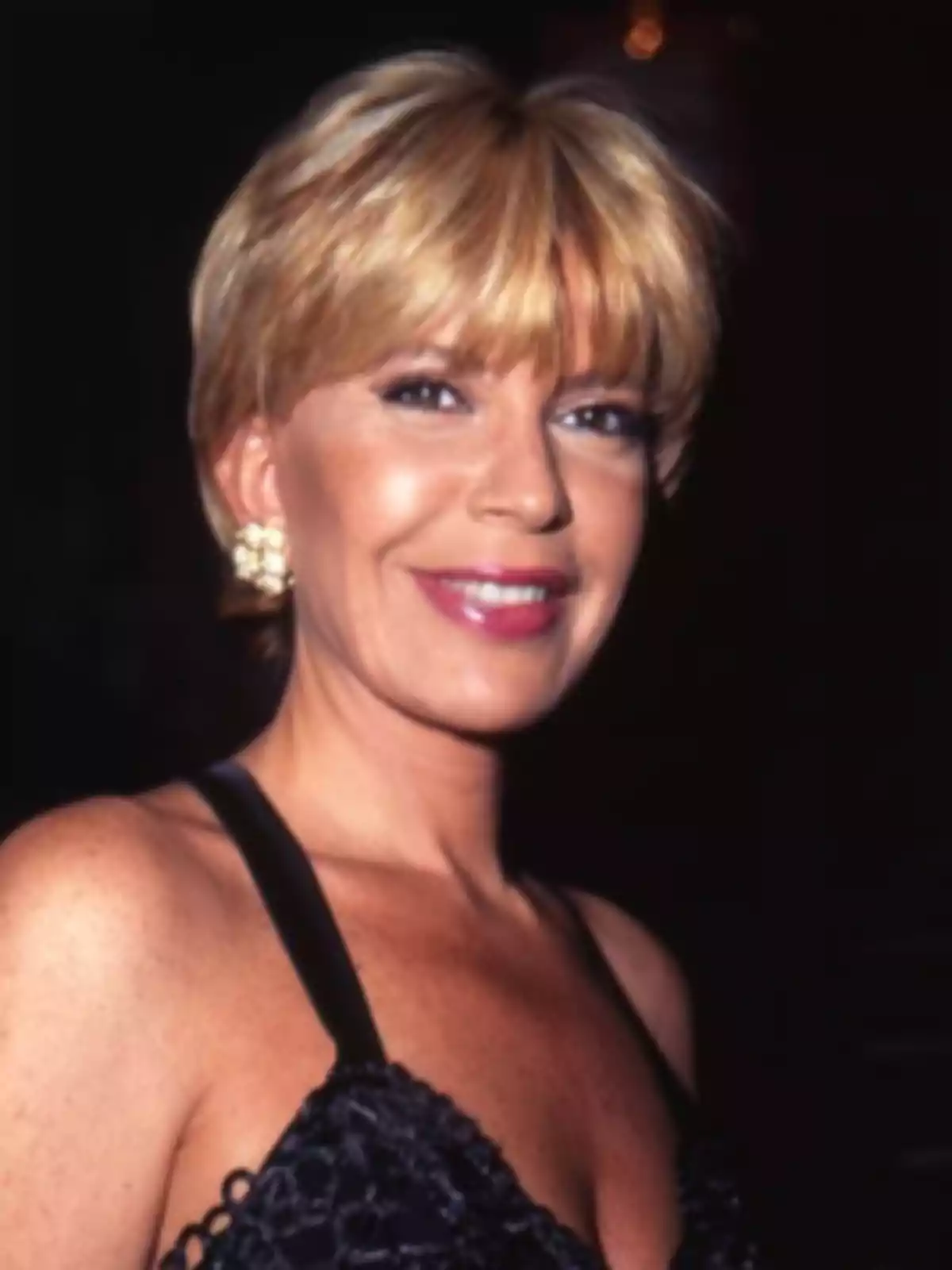 Mujer rubia con cabello corto y flequillo usando un vestido negro elegante y aretes dorados, sonriendo frente a un fondo oscuro Mujer rubia con cabello corto y flequillo usando un vestido negro elegante y aretes dorados, sonriendo frente a un fondo oscuro