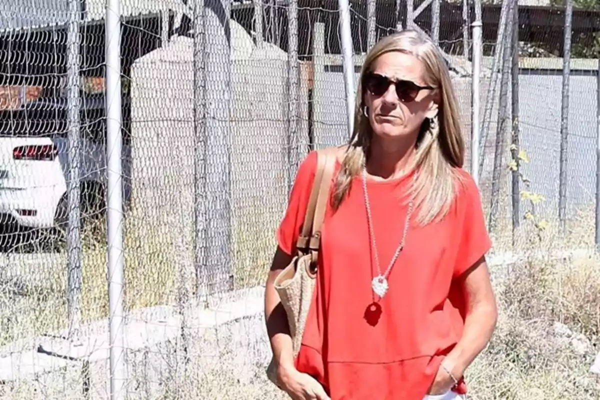 Mujer de cabello rubio y lacio usando gafas de sol, blusa roja y bolso beige, caminando al aire libre junto a una cerca de alambre Mujer de cabello rubio y lacio usando gafas de sol, blusa roja y bolso beige, caminando al aire libre junto a una cerca de alambre