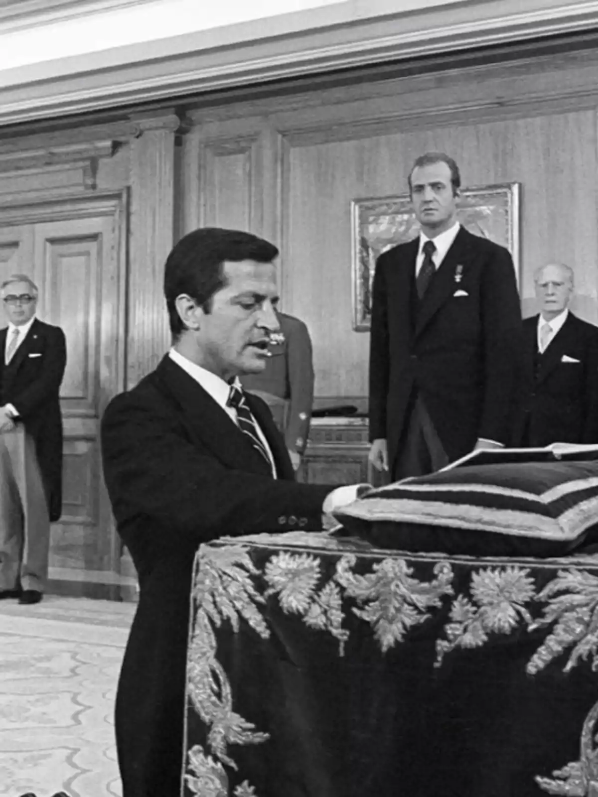 Un hombre de traje jura ante una mesa decorada mientras varias personas observan en una sala elegante Un hombre de traje jura ante una mesa decorada mientras varias personas observan en una sala elegante