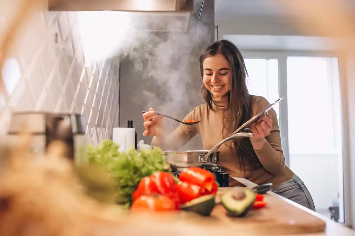 Una mujer sonriente cocina en una cocina moderna mientras prueba una sopa con una cuchara, rodeada de verduras frescas como pimientos y aguacate.