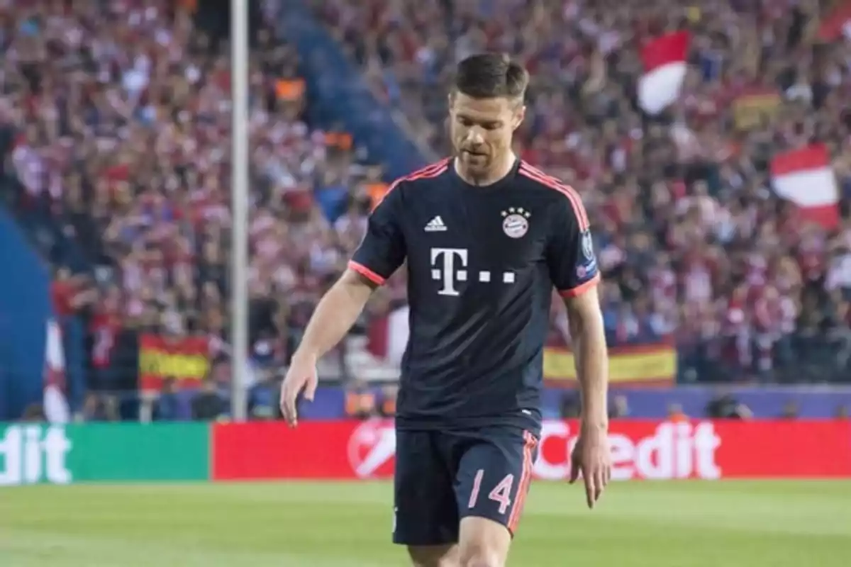 Jugador de fútbol con uniforme del Bayern Múnich caminando en el campo durante un partido con público en las gradas.