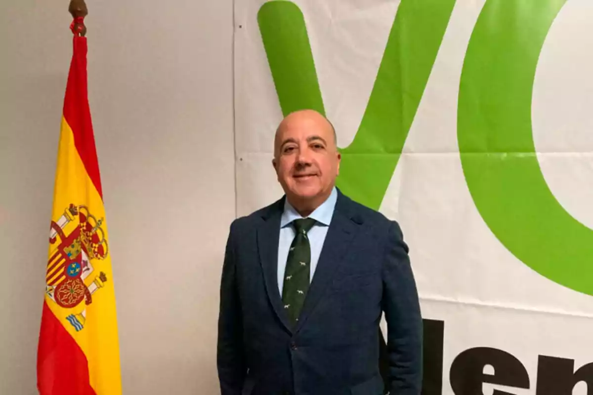 Hombre de traje posando junto a una bandera de España y un cartel con letras verdes.