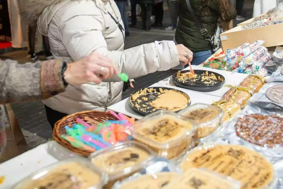 Personas probando dulces en un mercado al aire libre con una mesa llena de productos y utensilios de colores.