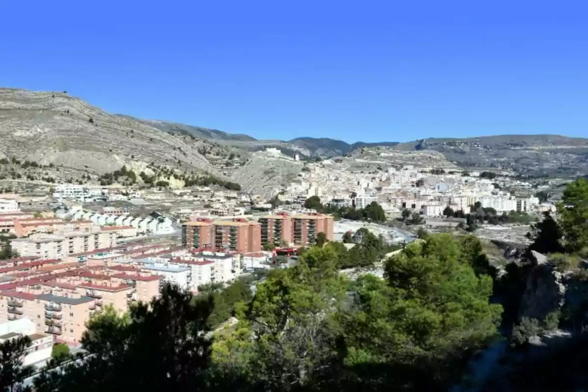 Vista panorámica de una ciudad rodeada de montañas con edificios y vegetación en primer plano bajo un cielo despejado.