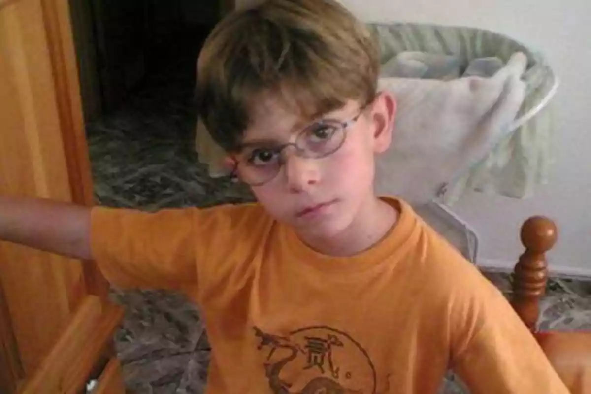 Un niño con gafas y camiseta naranja mira a la cámara mientras está en una habitación.