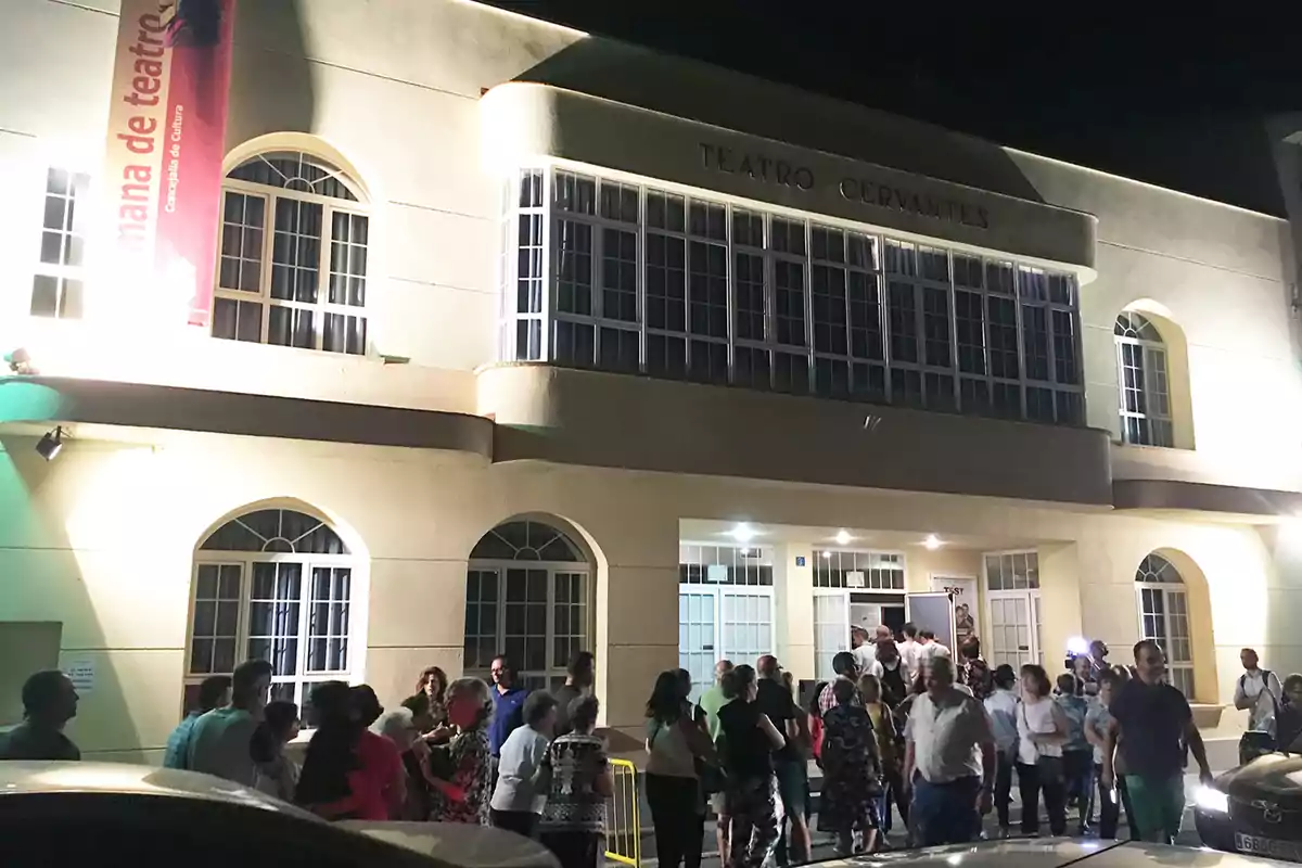 Un grupo de personas se reúne frente a la entrada iluminada del Teatro Cervantes durante la noche.