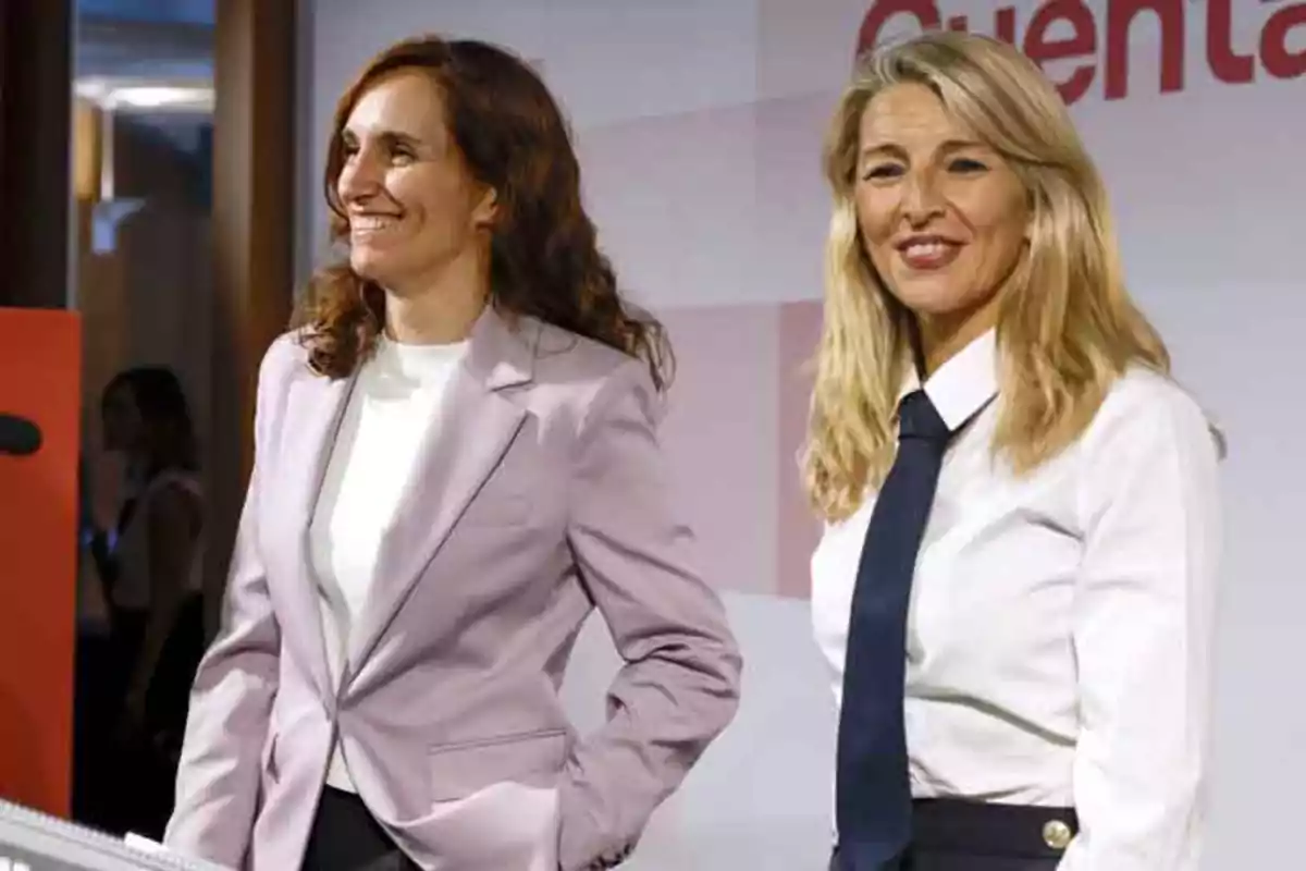 Dos mujeres sonrientes en un evento formal, una con chaqueta lila y la otra con camisa blanca y corbata negra.